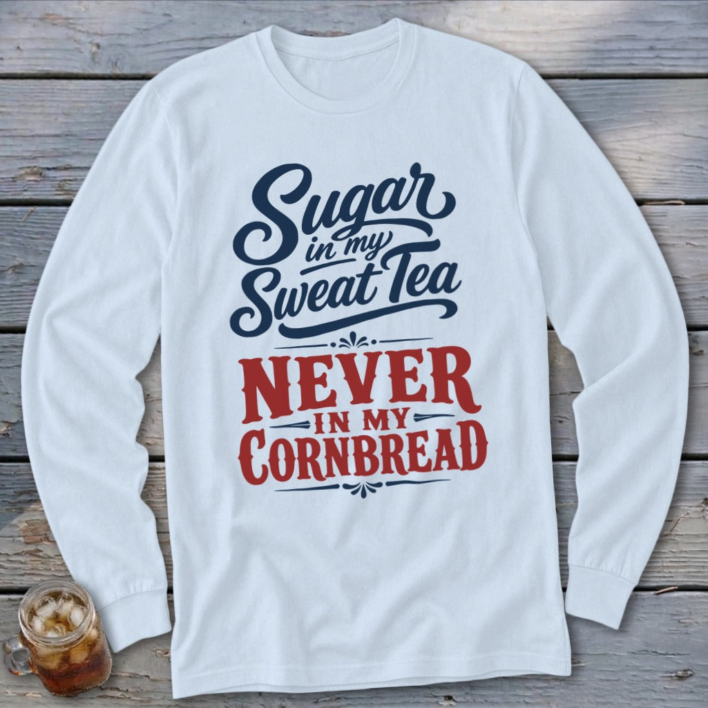 A white long-sleeved shirt with blue and red text reading Sugar in my Sweet Tea, Never in my Cornbread on a rustic wooden background.