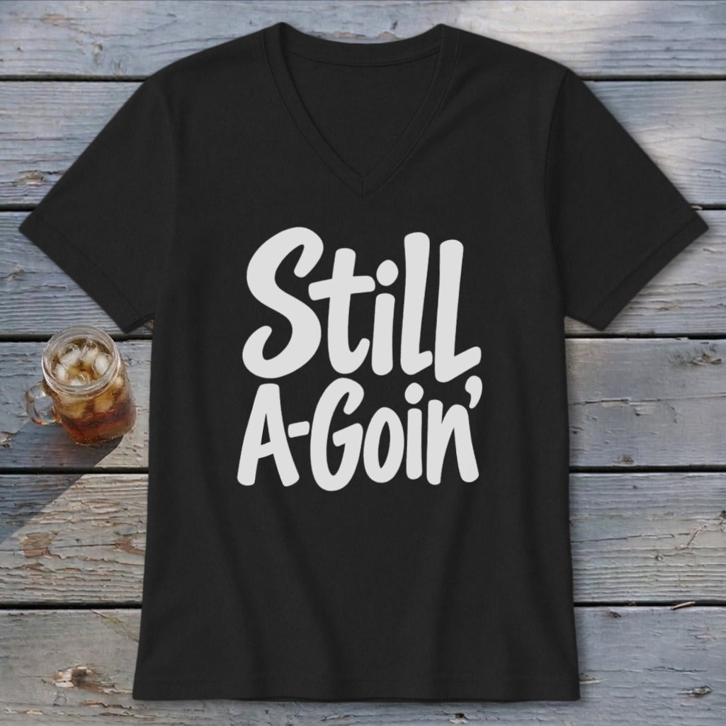 A black V-neck t-shirt with white text reading "Still A-Goin'" laid flat on a rustic wooden background.