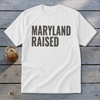 White t-shirt with dark gray text that reads "MARYLAND RAISED."