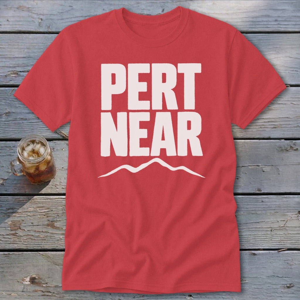 A red short-sleeve t-shirt with white block text reading PERT NEAR above a mountain outline, laid flat on a rustic wooden surface next to a glass of iced tea.