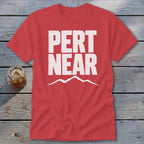 A red short-sleeve t-shirt with white block text reading PERT NEAR above a mountain outline, laid flat on a rustic wooden surface next to a glass of iced tea.