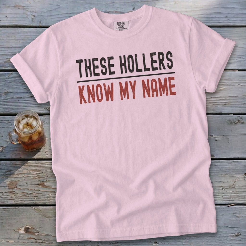 Light pink crewneck t-shirt with rolled sleeves laid flat on a rustic wooden surface, featuring black and red text reading THESE HOLLERS KNOW MY NAME.