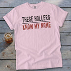 Light pink crewneck t-shirt with rolled sleeves laid flat on a rustic wooden surface, featuring black and red text reading THESE HOLLERS KNOW MY NAME.