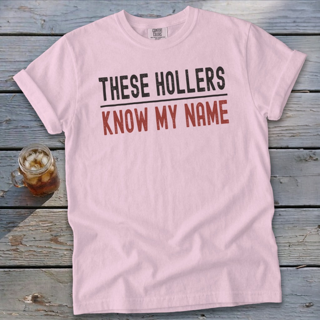 Light pink crewneck t-shirt with rolled sleeves laid flat on a rustic wooden surface, featuring black and red text reading THESE HOLLERS KNOW MY NAME.