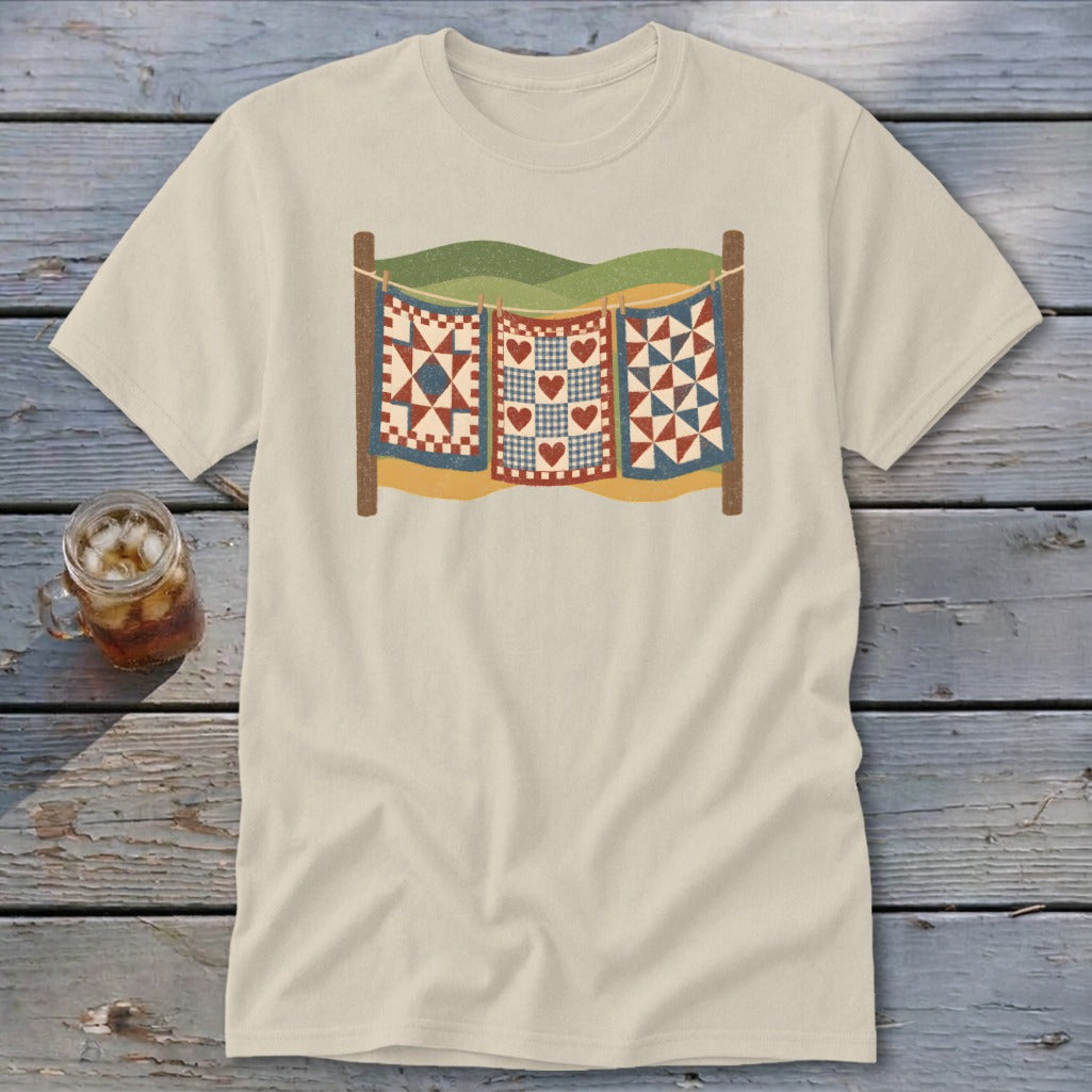 A cream-colored t-shirt with a vintage graphic of three quilts hanging on a clothesline.