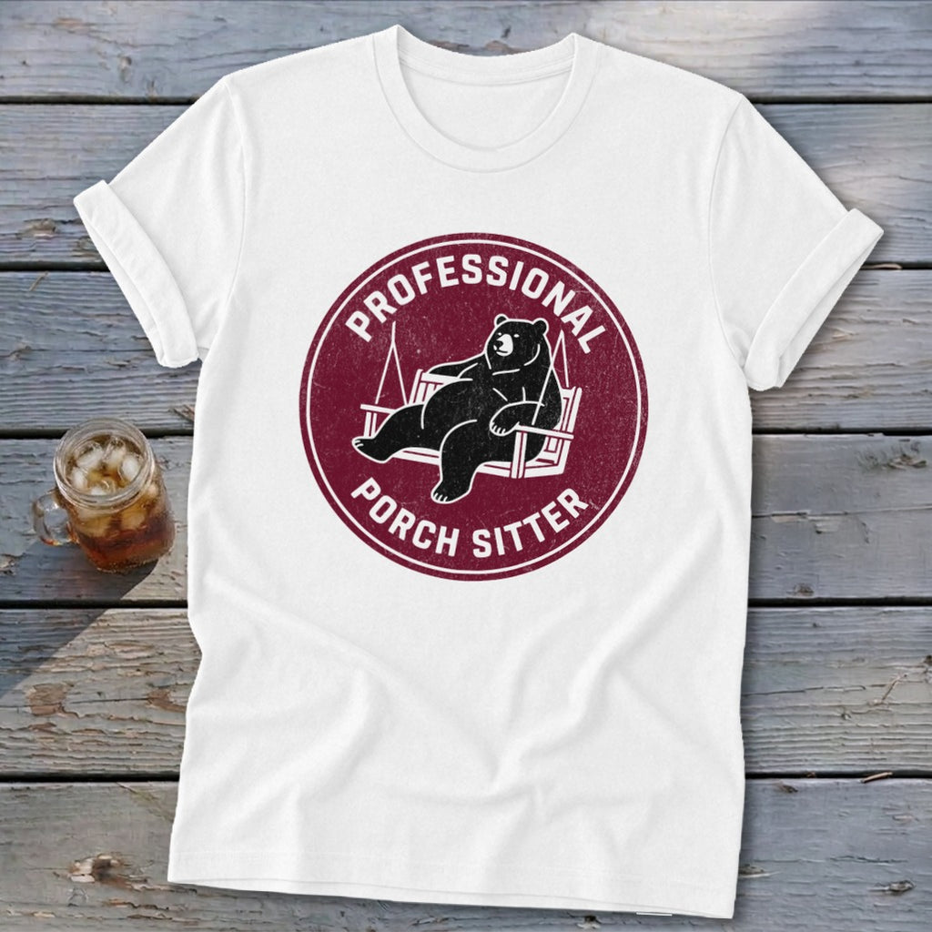 A white crewneck t-shirt featuring a maroon circular graphic with a black bear lounging on a porch swing and the text Professional Porch Sitter.
