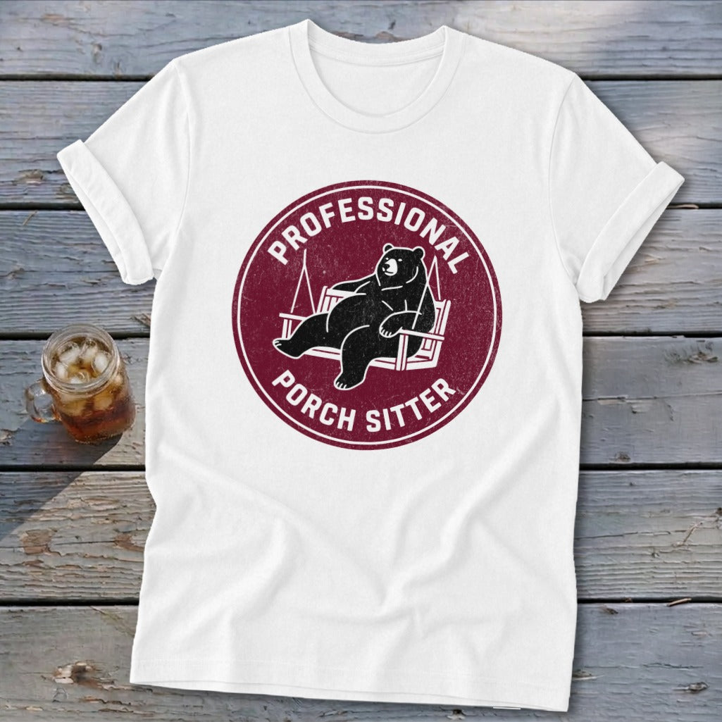 A white crewneck t-shirt featuring a maroon circular graphic with a black bear lounging on a porch swing and the text Professional Porch Sitter.