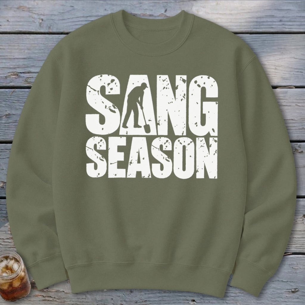 An olive green crewneck sweatshirt featuring distressed white text that reads SANG SEASON, with the silhouette of a digger holding a shovel inside the letter A.