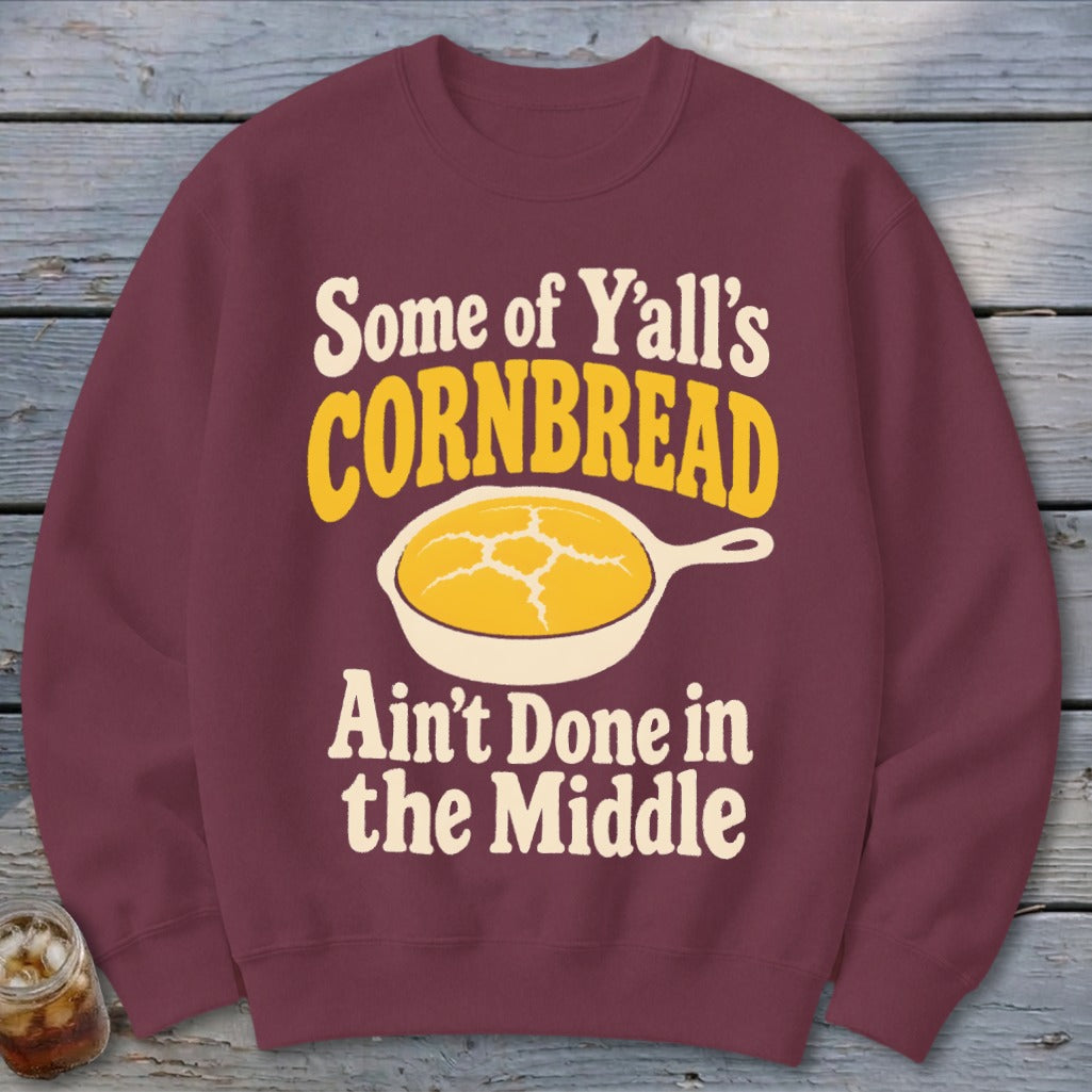 A maroon crewneck sweatshirt with graphic text reading Some of Y'all's Cornbread Ain't Done in the Middle above a skillet of cornbread.