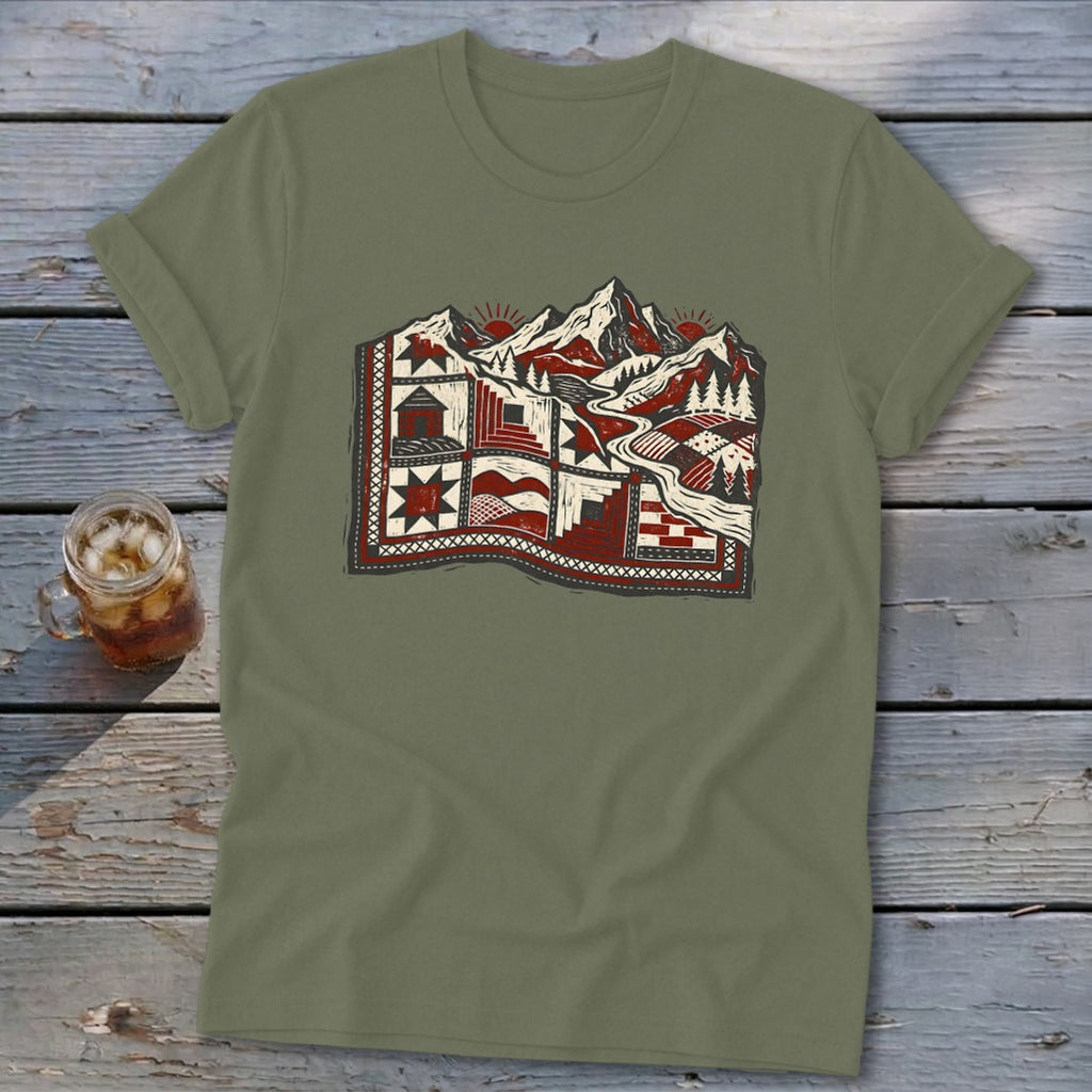 An olive green t-shirt featuring a stylized graphic of snowy mountains and a patchwork quilt on a wooden surface.
