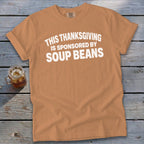 Tan t-shirt with white text reading "This Thanksgiving is sponsored by soup beans" laid flat on a rustic wooden surface.