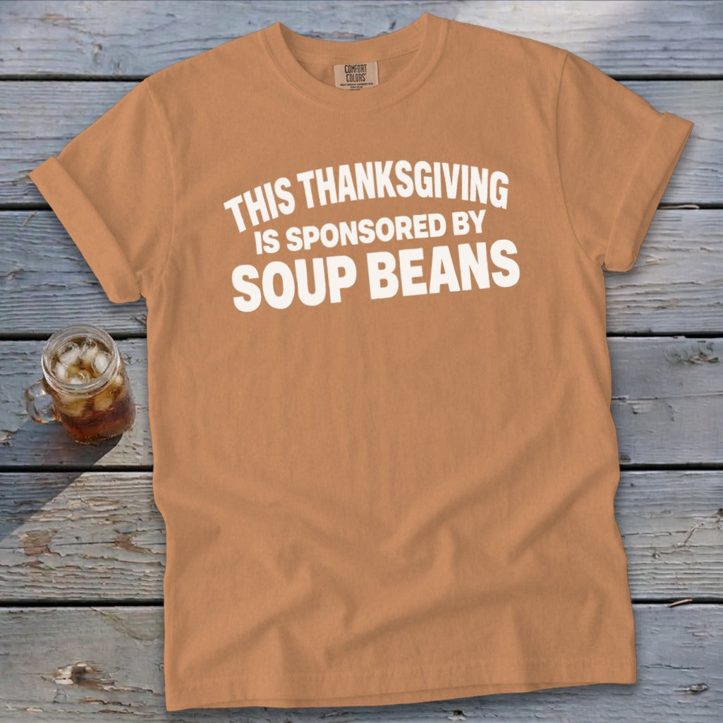 Tan t-shirt with white text reading "This Thanksgiving is sponsored by soup beans" laid flat on a rustic wooden surface.