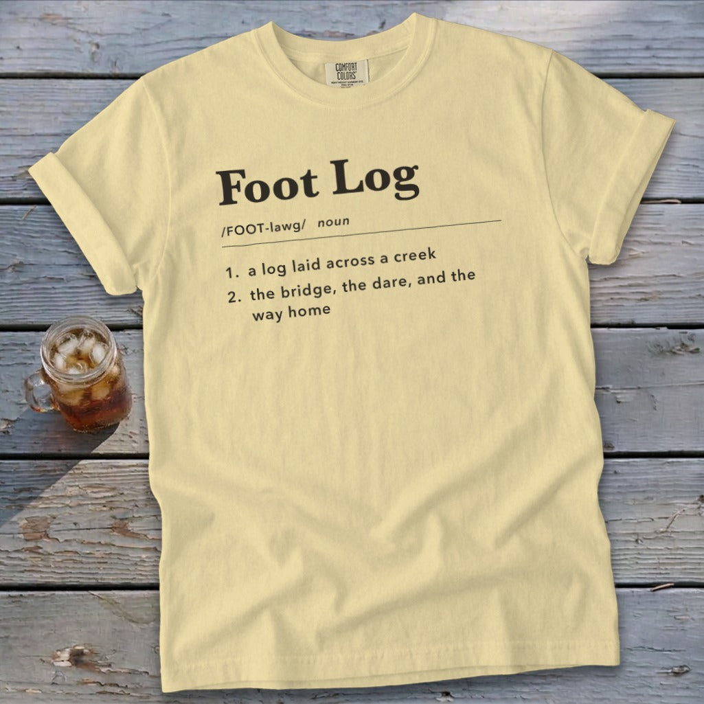 A pale yellow t-shirt with black dictionary-style text defining Foot Log lies on a wooden surface next to an iced drink.