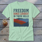 A light green t-shirt featuring the text FREEDOM RINGS LOUDEST IN THESE HILLS above a red, blue, and dark blue mountain landscape graphic.