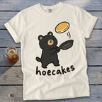An off-white t-shirt featuring a cartoon black bear flipping a pancake in a skillet with the text hoecakes underneath.