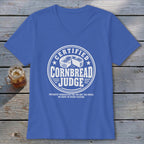 A blue V-neck t-shirt with a white graphic reading Certified Cornbread Judge laid flat on weathered wood.