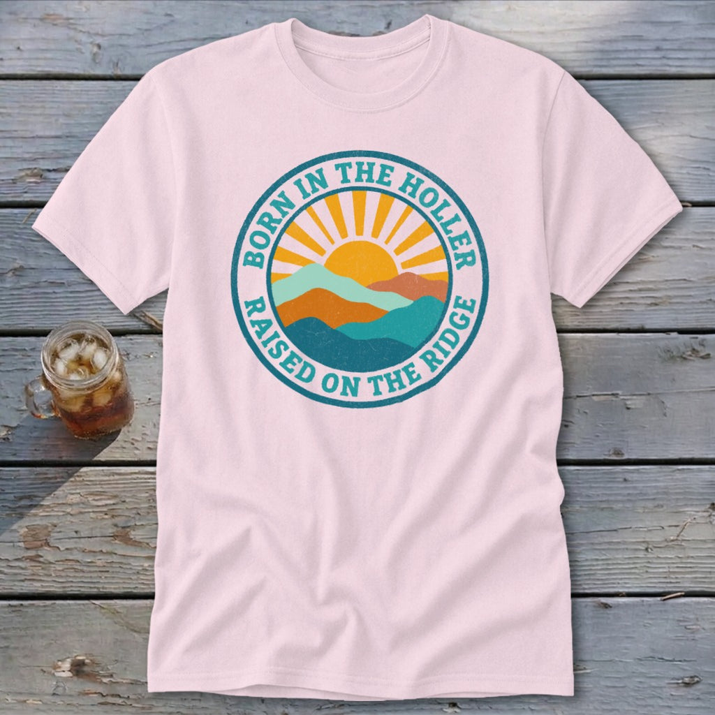 Light pink short-sleeve t-shirt with a round graphic of mountains, a sun, and the text Born in the Holler Raised on the Ridge.