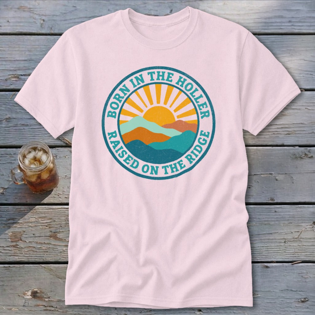 Light pink short-sleeve t-shirt with a round graphic of mountains, a sun, and the text Born in the Holler Raised on the Ridge.