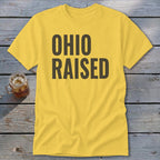 Yellow t-shirt with "OHIO RAISED" text in dark gray, next to a glass of iced tea.