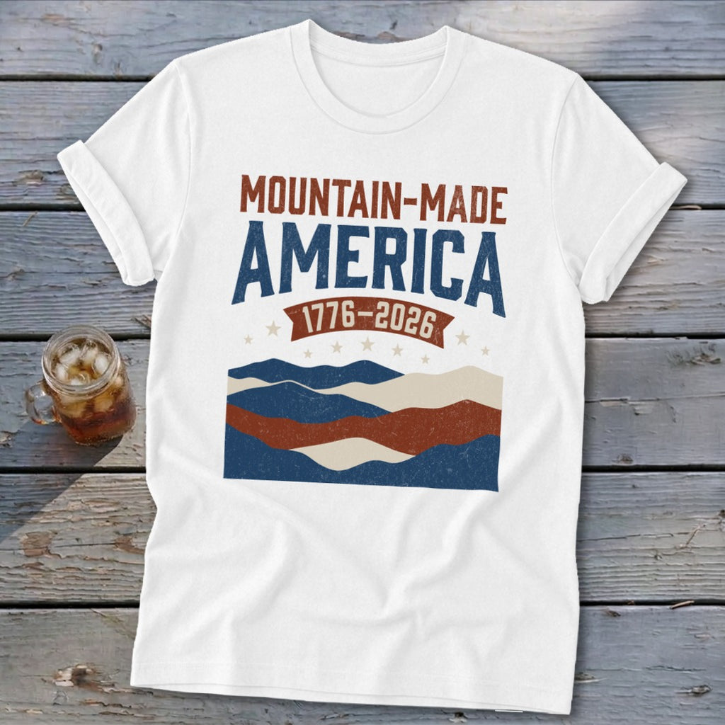 A white short-sleeved t-shirt featuring a distressed red, white, and blue graphic with text that reads Mountain-Made America 1776-2026.