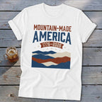 A white short-sleeved t-shirt featuring a distressed red, white, and blue graphic with text that reads Mountain-Made America 1776-2026.