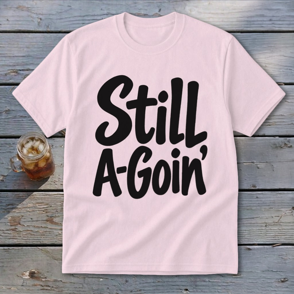 Light pink t-shirt featuring the black text "Still A-Goin'" laid flat on weathered wooden boards.