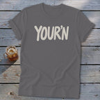 Gray t-shirt with "YOUR'N" text and a drink on a wooden background.