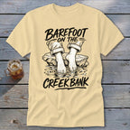 Light yellow t-shirt with "Barefoot on the Creek Bank" text and illustration of feet in water.