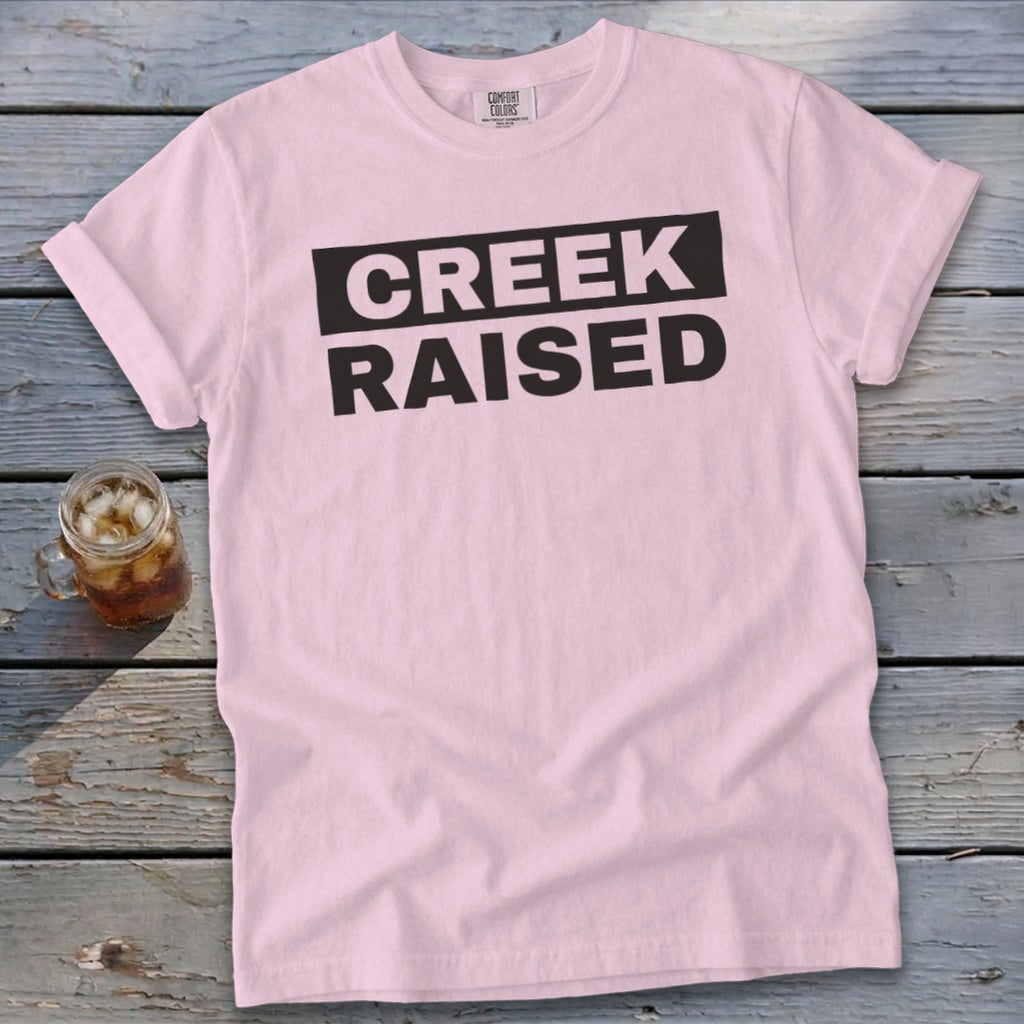 Pink Comfort Colors t-shirt with black text reading "CREEK RAISED".