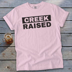 Pink Comfort Colors t-shirt with black text reading "CREEK RAISED".
