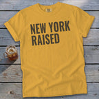 Yellow t-shirt with "NEW YORK RAISED" text on a wooden background.