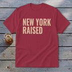 Red t-shirt with tan text reading "New York Raised" on a wooden surface.