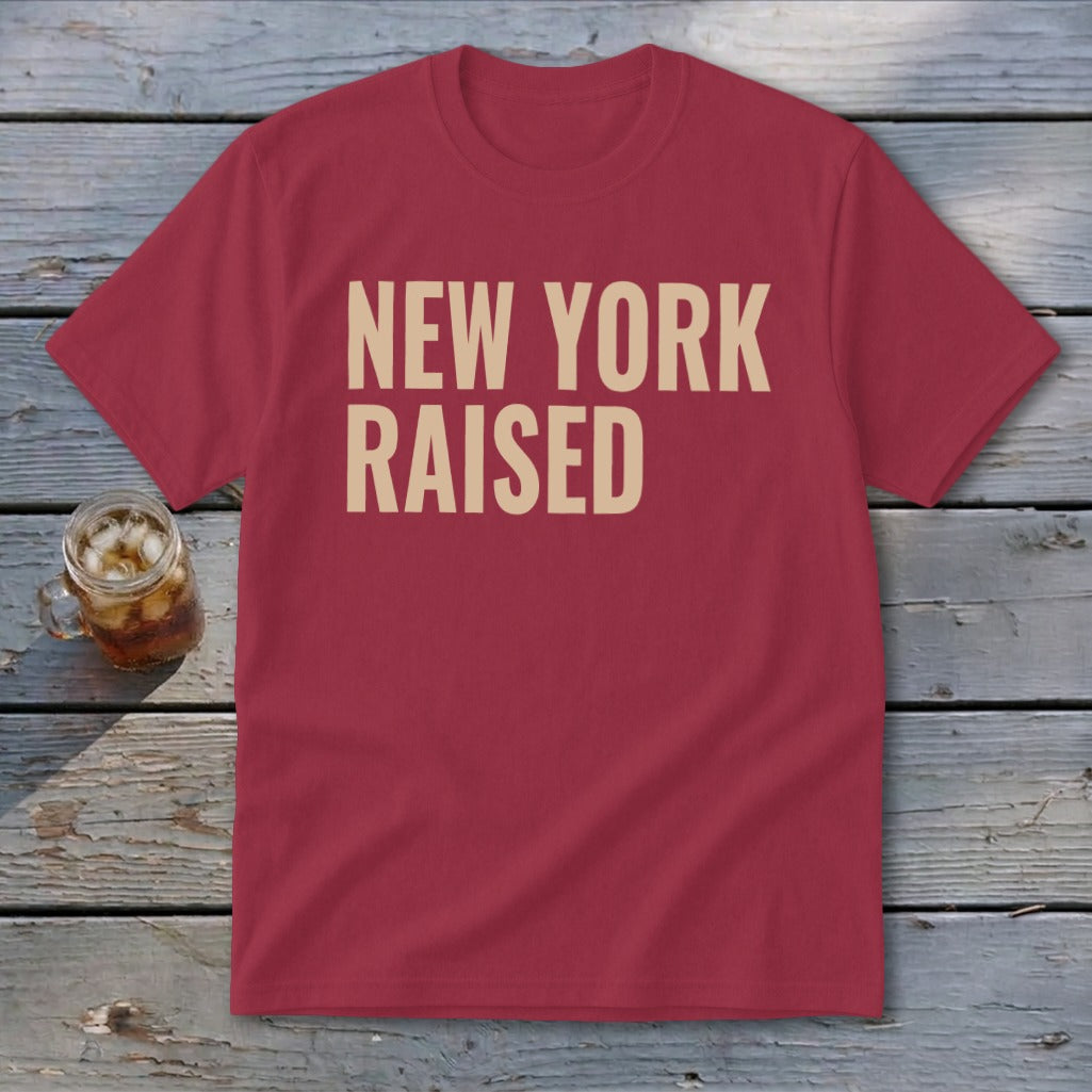 Red t-shirt with tan text reading "New York Raised" on a wooden surface.