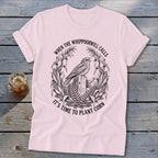 Light pink t-shirt featuring a black line art graphic of a whippoorwill, corn stalks, and text.
