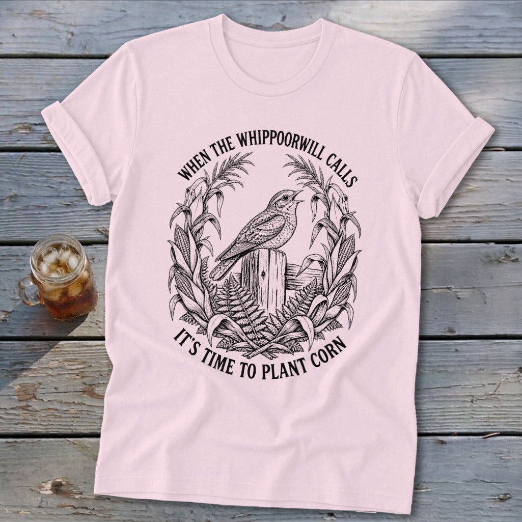 Light pink t-shirt featuring a black line art graphic of a whippoorwill, corn stalks, and text.