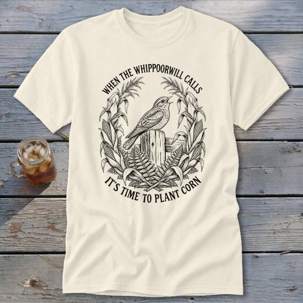 A cream t-shirt with a black line art graphic of a whippoorwill, corn stalks, and text.