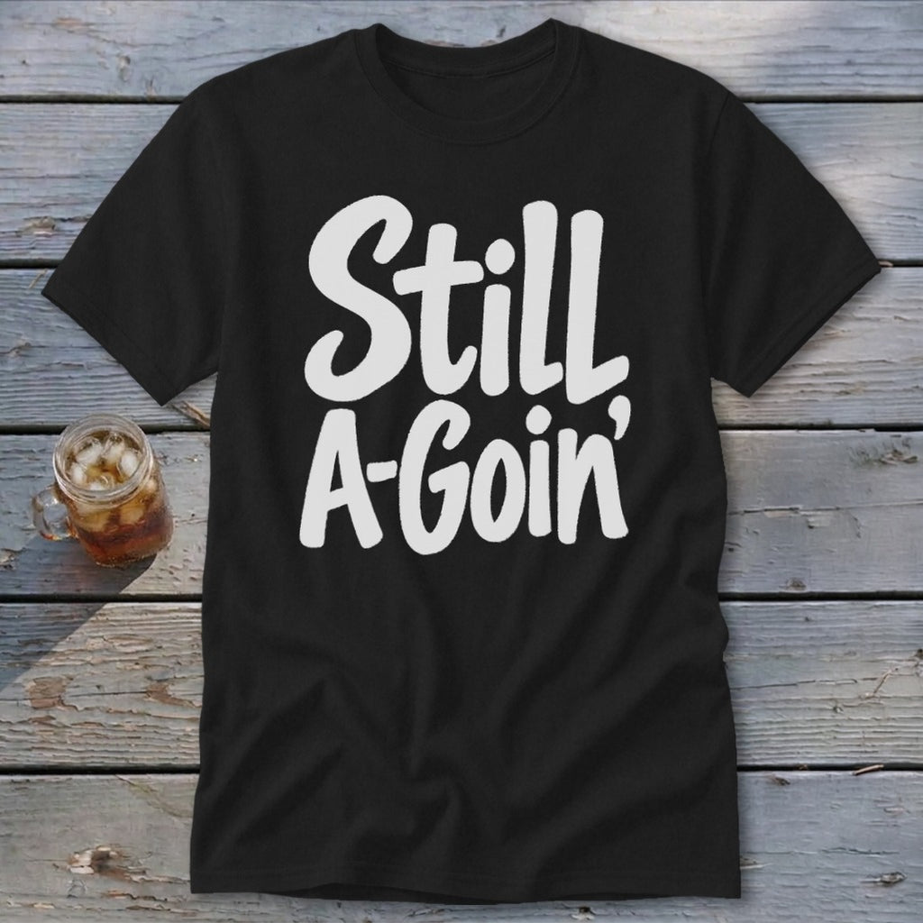 Black short-sleeved t-shirt with white handwritten-style text reading Still A-Goin' on a wooden surface.