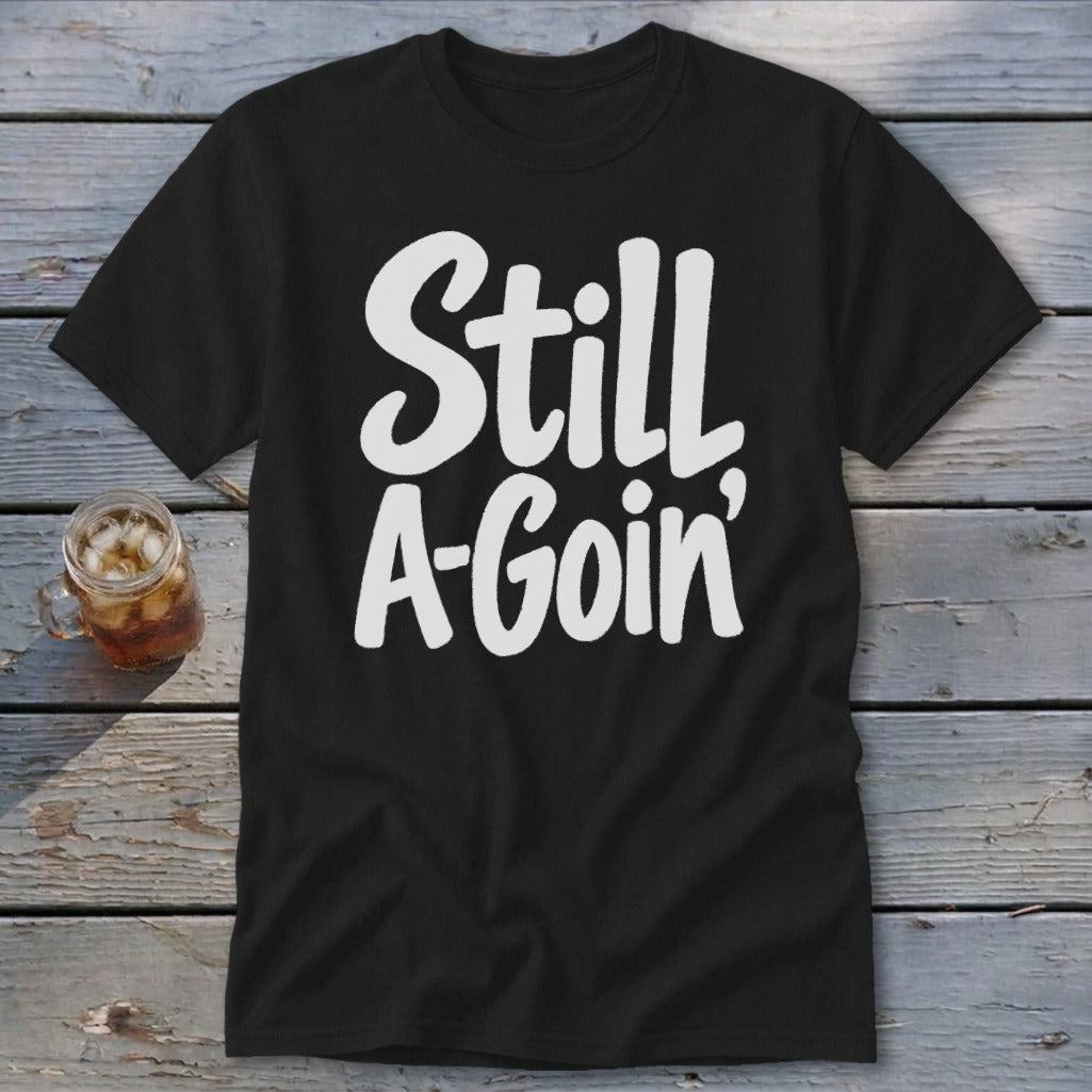 Black short-sleeved t-shirt with white handwritten-style text reading Still A-Goin' on a wooden surface.