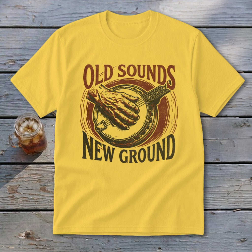 Yellow t-shirt with a vintage banjo design and the text OLD SOUNDS NEW GROUND, laid flat on a rustic wooden table.