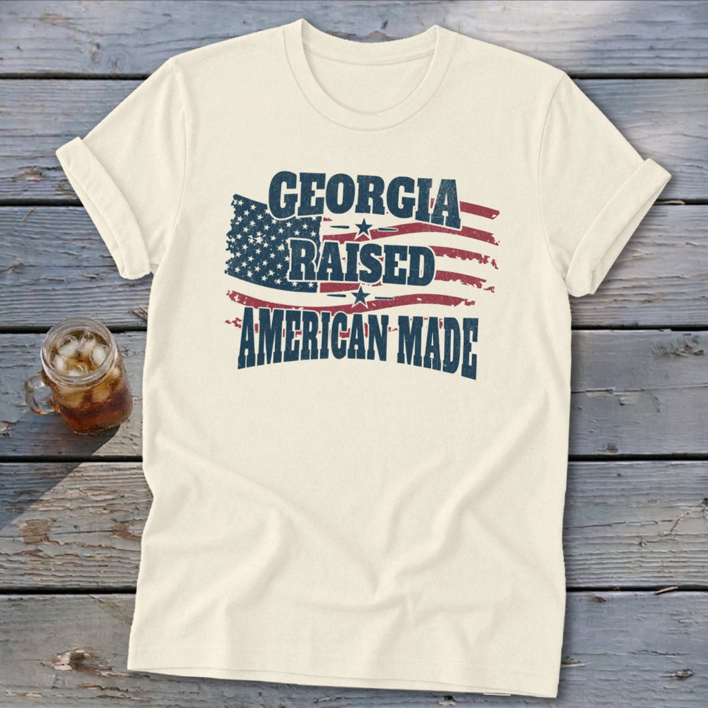 An off-white t-shirt with a distressed graphic of an American flag and the text Georgia Raised American Made.