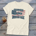 An off-white t-shirt with a distressed graphic of an American flag and the text Georgia Raised American Made.
