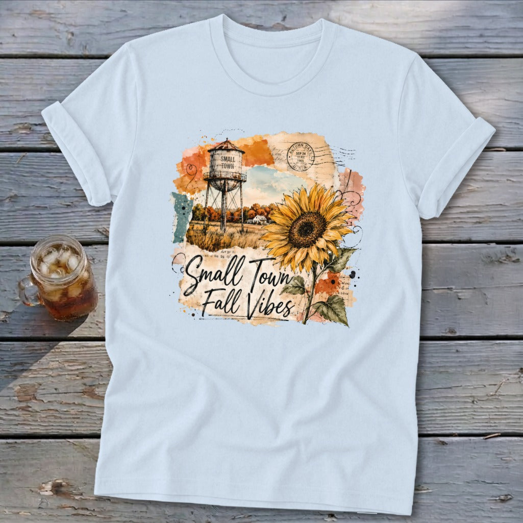 Light blue t-shirt with a graphic featuring a water tower, sunflower, and text "Small Town Fall Vibes."
