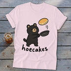 Light pink t-shirt featuring a cartoon black bear flipping a pancake from a skillet, with the text hoecakes.