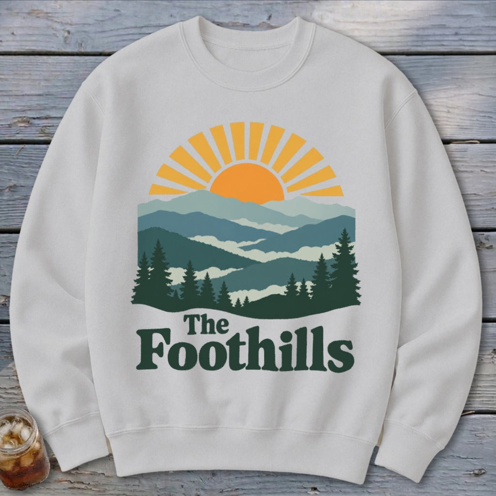 White sweatshirt with sun and mountain graphic and "The Foothills" text.