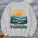 White sweatshirt with sun and mountain graphic and "The Foothills" text.