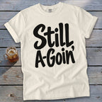 An off-white short-sleeve t-shirt with black script text reading Still A-Goin' on a wooden surface.