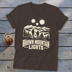 A brown short-sleeve t-shirt with a graphic of three figures, mountains, and glowing lights.