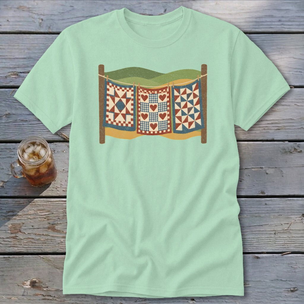 A light green t-shirt featuring a graphic of three patchwork quilts hanging on a clothesline.