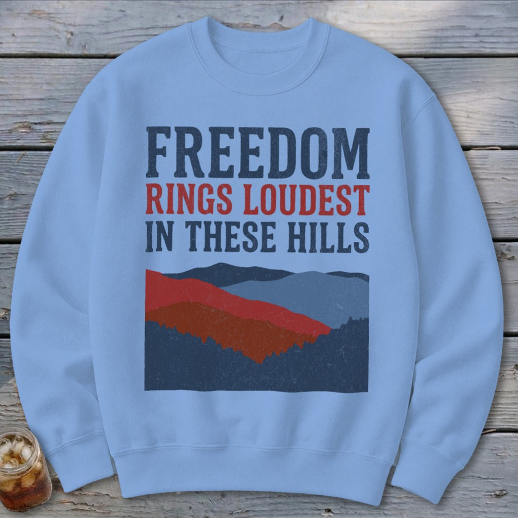 Light blue crewneck sweatshirt featuring the text Freedom Rings Loudest in These Hills above layered mountain graphics.