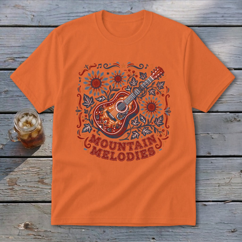 Orange t-shirt featuring a folk-style illustration of an acoustic guitar, sunflowers, leaves, and the text MOUNTAIN MELODIES.